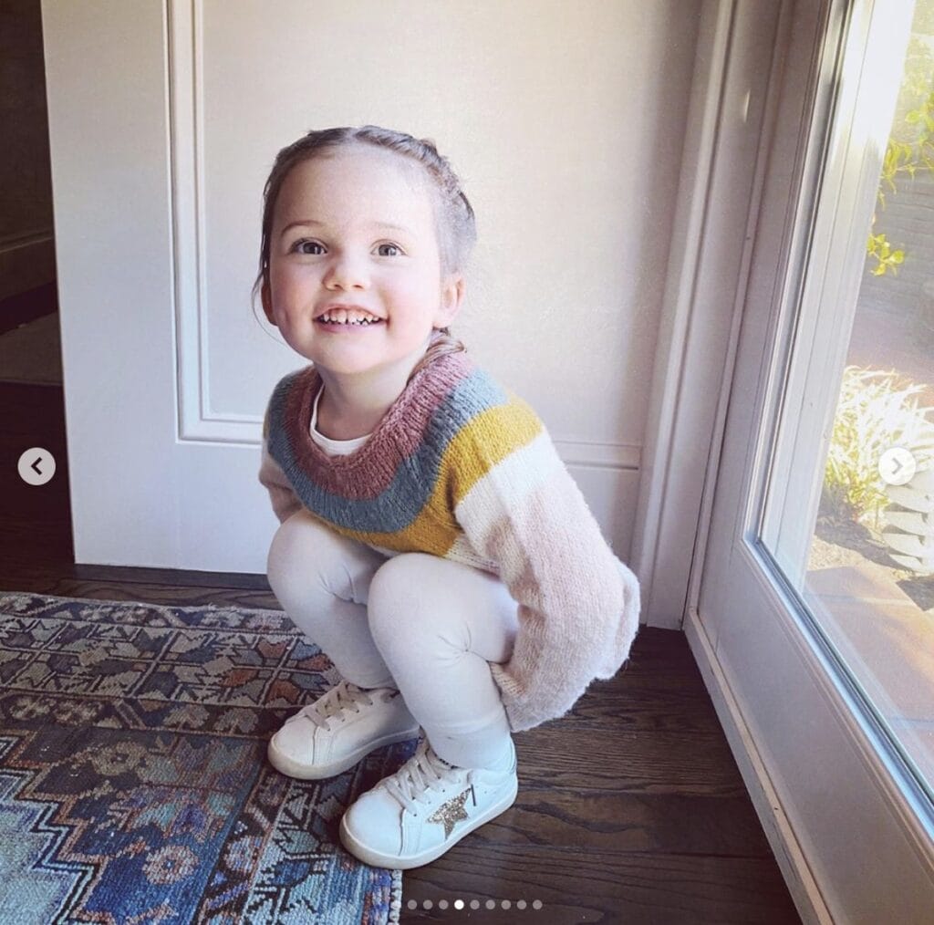 Young girl squats inside home while smiling directly at the camera. She is wearing white pants & shoes with a striped sweater. 