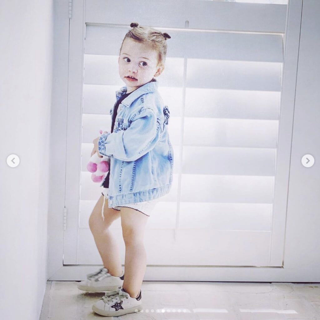 Young girl standing in home looks off camera while holding a pink and white stuffed animal. She wears shorts and sports an oversized jean jacket