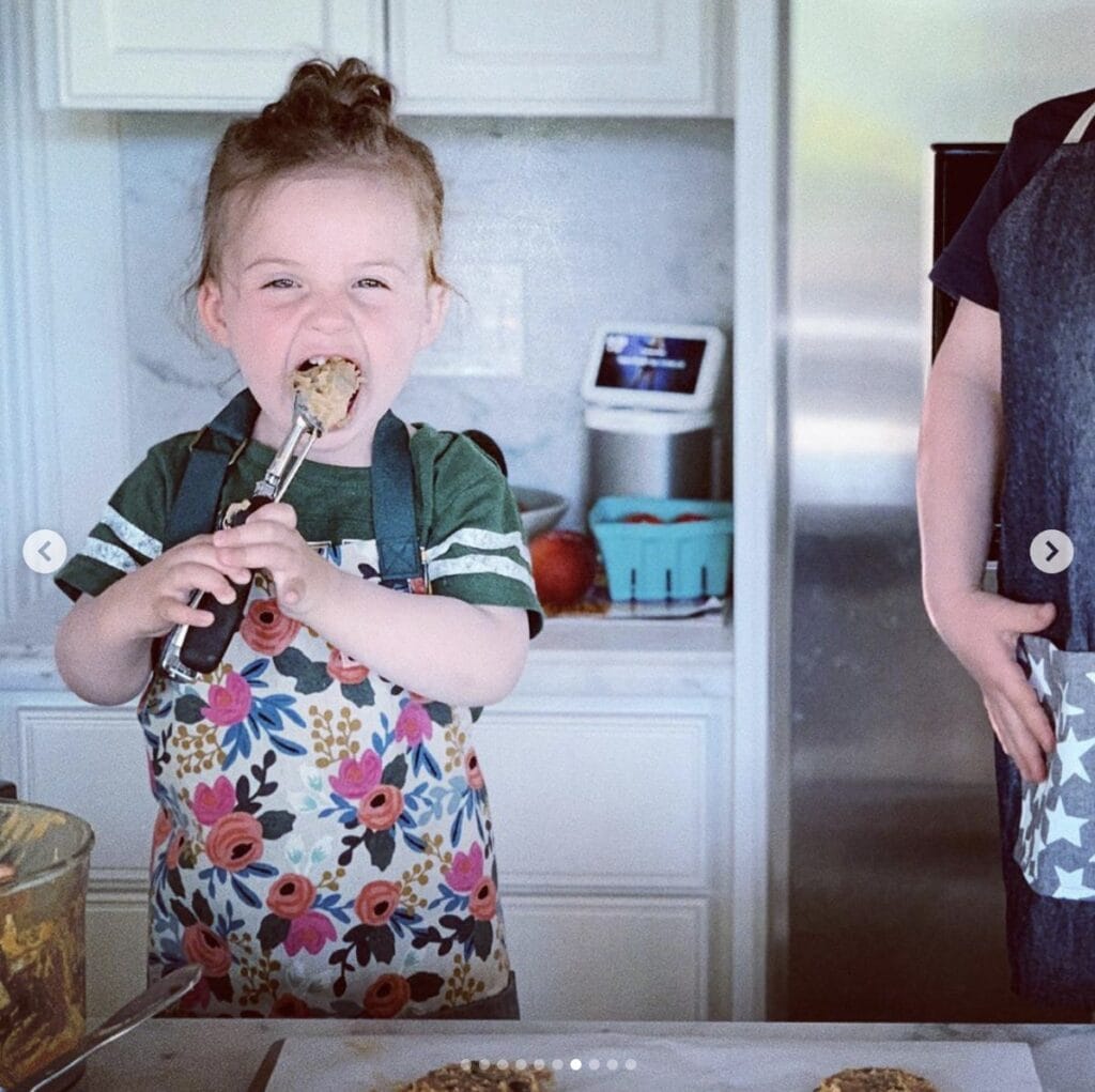 Young girl stands in home kitchen wearing a floral apron and enthusiastically licks batter off a spoon