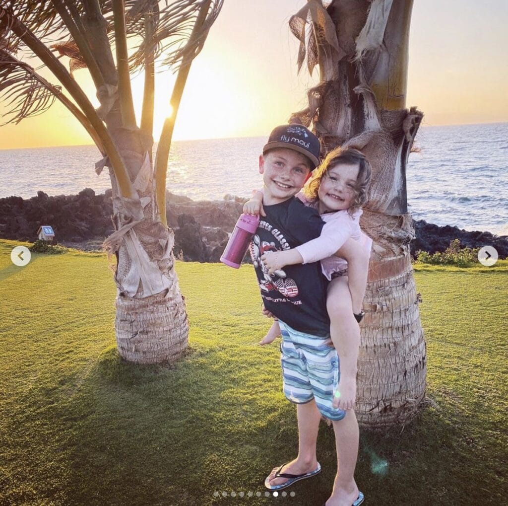 young girl is given a piggyback ride by her older brother. They both smile at the camera while they stand on a lawn that features a sunset over the ocean