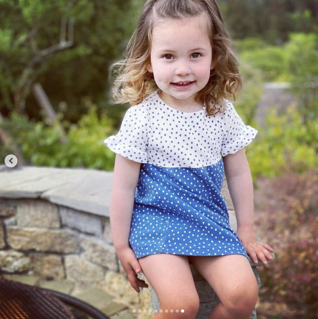 young girl smiles directly at camera wearing a blue and white polka dot dress with ruffled short sleeves