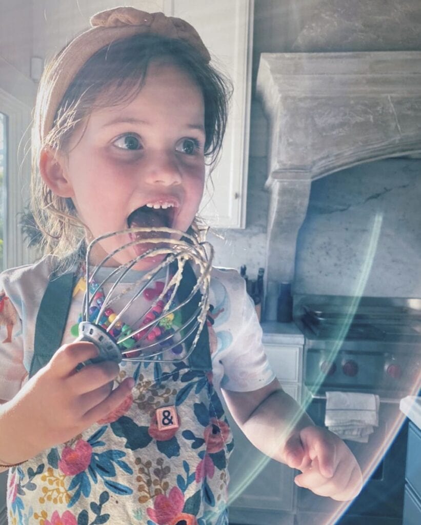 Young girl in home kitchen wears a floral apron and licks batter off a whisk