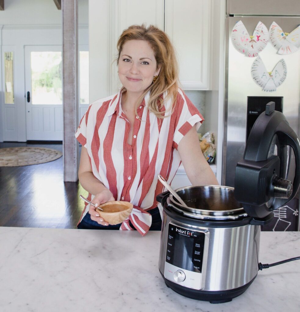 Woman wearing striped shirt stands in home kitchen next to Instant Pot holding a bowl of fresh homemade applesauce 