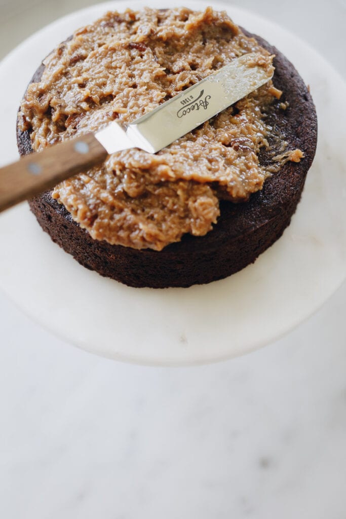 German Chocolate Cake in the middle of being frosted as it rests on a white cake stand