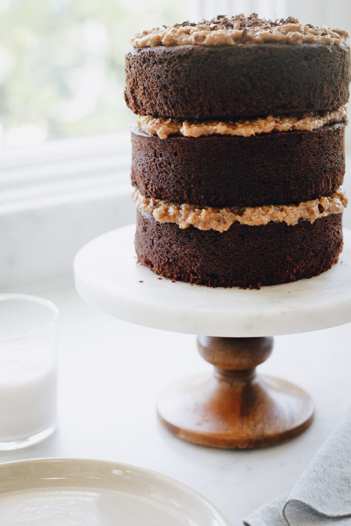 3 layer Gluten Free German Chocolate Cake resting on a cake stand by a window. A glass of milk is next to it. 