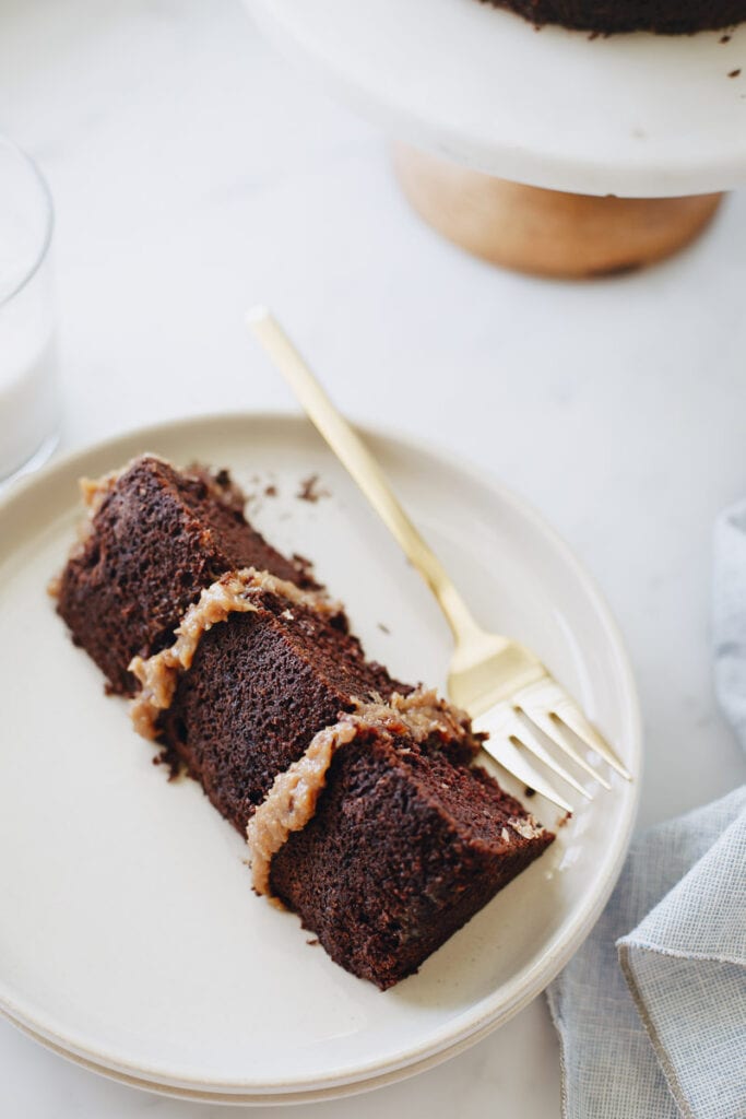 slice of Gluten Free German Chocolate Cake on a white plate with a gold colored fork next to it