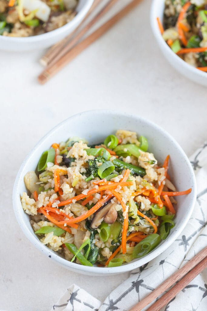 veggie fried rice in a white bowl with a pair of wooden chopsticks laying on a cloth napkin to it's right 