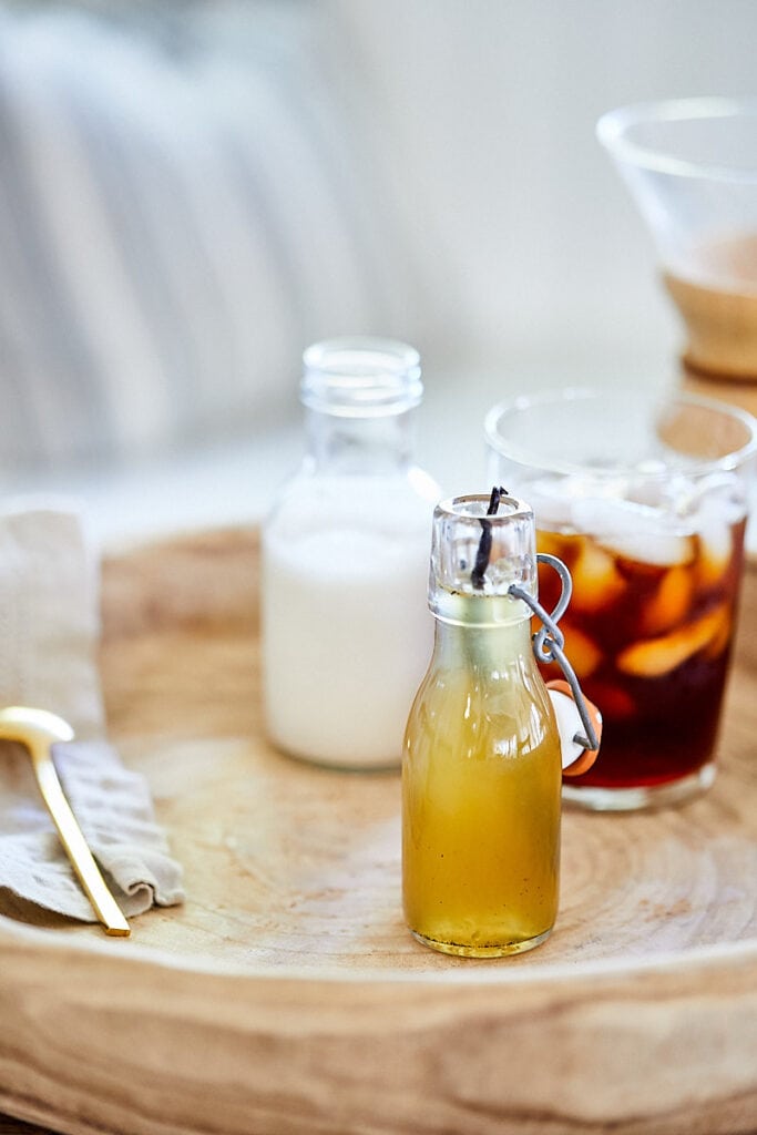 Vanilla Honey Simple Syrup in a glass bottle. Creamer and an iced coffee are in the background. 