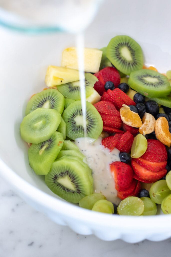 dairy free yogurt being poured over an assortment of fruit such as kiwi, strawberries, pineapple, blueberries and mandarins