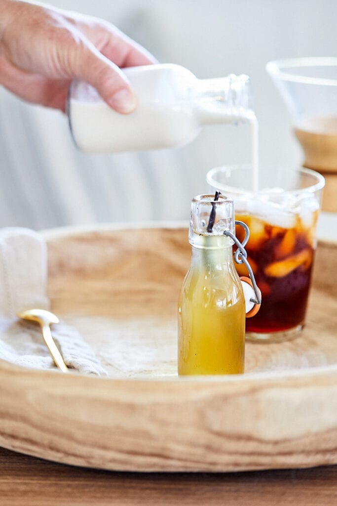 Vanilla Honey Simple Syrup in a glass bottle on a wooden tray. A hand pours creamer into an iced coffee in the background.