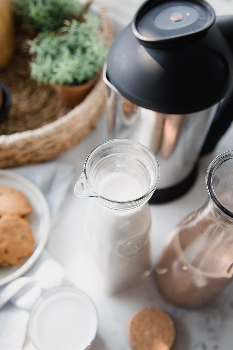  homemade dairy-free milk in glass carafes on a table next to a plate with chocolate chips